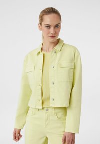 Jasnożółta kurtka dżinsowa z dwoma przednimi kieszeniami, zapinana na napy, z długimi rękawami. Noszona na pasującym jasnożółtym topie.