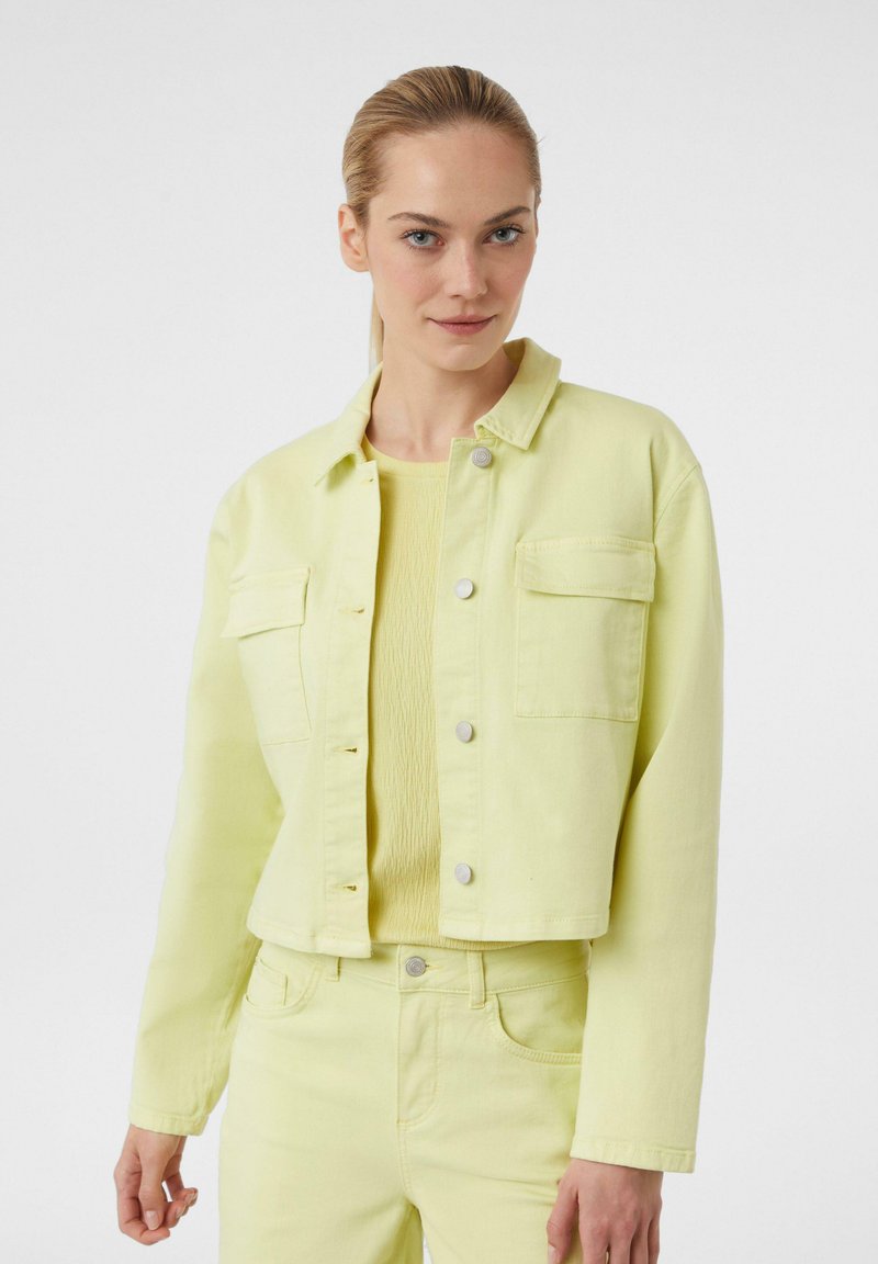 Jasnożółta kurtka dżinsowa z dwoma przednimi kieszeniami, zapinana na napy, z długimi rękawami. Noszona na pasującym jasnożółtym topie.