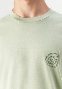 T-shirt en coton vert clair avec un col rond, arborant un petit design en spirale dans une teinte plus foncée. Texture lisse, manches courtes.
