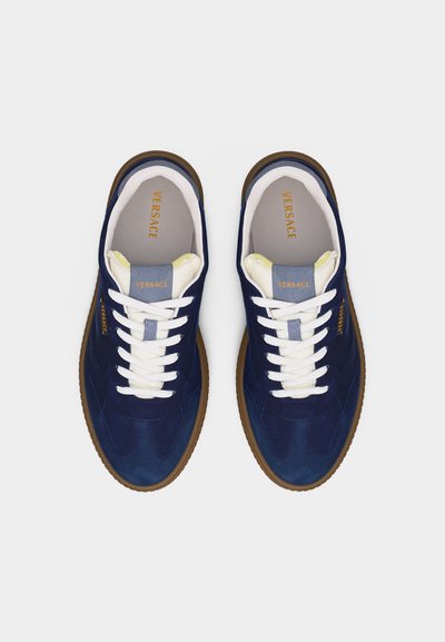 Paar dunkelblaue Versace-Sneaker mit weißen Schnürsenkeln, hellbeigen Innensohlen und braunen Gummisohlen, von oben auf weißem Hintergrund betrachtet.