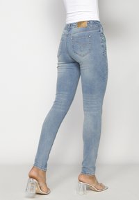 Jeans ajustados en denim azul claro con un acabado deslavado, que presentan un diseño de bolsillo en la parte trasera y detalles a lo largo del lateral. Sandalias con tacones transparentes.