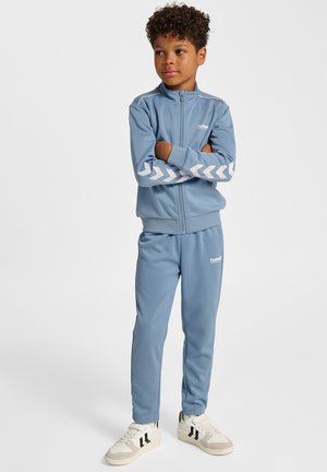 Lichtblauw trainingspak van gladde stof, met een jack met ritssluiting en witte chevron-accenten op de mouwen, en bijpassende broek.