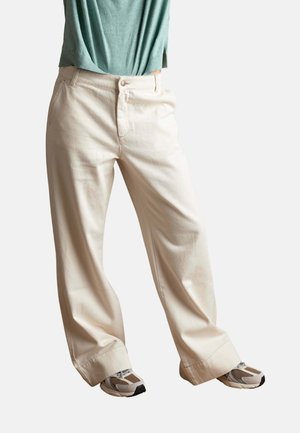 Persona que lleva puestos pantalones sueltos color marfil con puños anchos y zapatillas beige, sobre un fondo blanco liso.