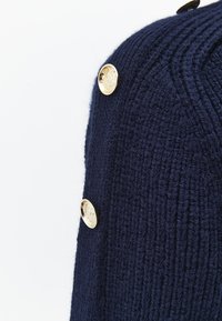 Marineblauer Strickpullover mit geripptem Muster, figurbetontem Schnitt und goldfarbenen Zierknöpfen an der Seite.