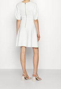 Bílé šaty s krátkým rukávem, s nabíranými rukávy, těsným topem, rozšířenou sukní, zadním zipem a hladkým povrchem. V kombinaci s béžovými sandály na blokovém podpatku.