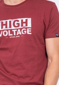 T-shirt court à manches courtes bordeaux avec un imprimé graphique blanc "HAUTE TENSION". Matière en coton doux, coupe décontractée, dotée d'une petite étiquette de marque sur la manche.