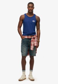 Marine tanktop met logo, denimshorts, rood-witte geruite shirt geknoopt om de taille, crèmekleurige sneakers en crèmekleurige sokken met blauwe strepen.