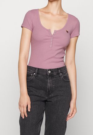 Osoba w krótkim rękawie, mauve (fioletowa) henley bluzce z guzikami i czarnych dżinsach, stoi na tle jasnego, jednolitego tła.
