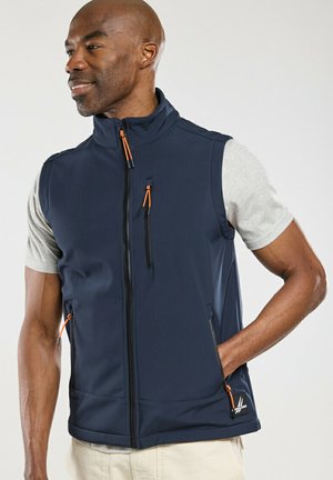 Homme portant un gilet zippé bleu marine avec des tirettes de fermeture éclair orange, par-dessus un t-shirt à manches courtes gris clair, main dans la poche, regardant vers la gauche.