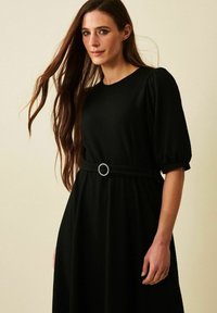 Musta mekko, jossa on teksturoitu kangas, lyhyet puffihihat, pyöreä hopeinen vyösolki ja liehuva hame. Minimalistinen muotoilu ilman kuvioita.