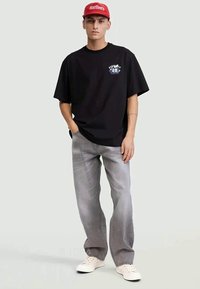 Jovem de pé, usando um boné vermelho, uma t-shirt gráfica preta, jeans cinzentos e ténis brancos em frente a um fundo liso claro.