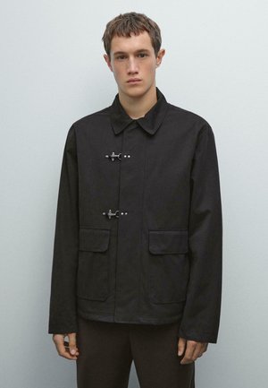 Black jacket featuring a button-up front with silver hardware, large patch pockets, and a classic collar. Smooth fabric with a slightly structured fit.