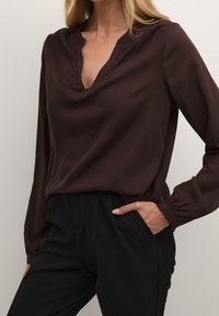 Brun langærmet bluse med V-hals og samlede manchetter, parret med sorte højhalsede bukser. Glat stoftekstur, minimalistisk design.