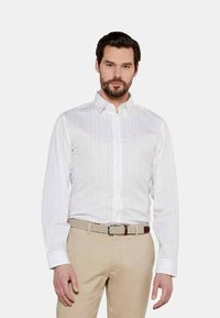 Camicia bianca con righe verticali rosse, maniche lunghe e colletto classico, abbinata a pantaloni beige e una cintura intrecciata.