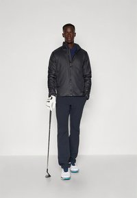 Black zip-up jacket, dark blue shirt, navy pants, white golf glove, and golf club. Shoes have white base with blue accents. Simple design.