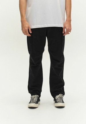 Person iført sorte cargobukser, sort-hvide høje sneakers og en hvid t-shirt, stående mod en ensfarvet baggrund.