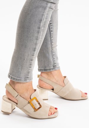 Beige suede block-heeled sandals with a clear acrylic buckle detail, open-toe design, and slingback strap; paired with gray denim.
