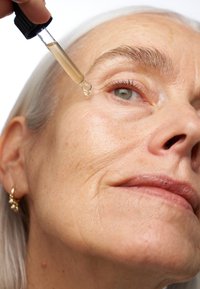 Clear serum dropper held above a face with light skin. The skin shows fine lines; hair is straight and silver. The background is white.