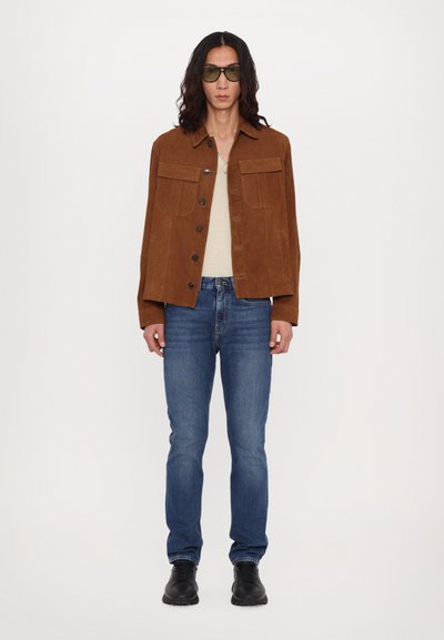 Veste en daim marron avec deux poches poitrine, chemise beige, jeans bleus et bottes noires ; le modèle se tient devant un fond blanc.