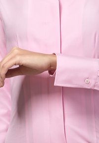 Camicia button-up rosa chiaro con righe verticali; maniche lunghe; polsino visibile con bottone; texture del tessuto liscia e morbida.