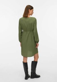 Sieviete, tērpta olīvzaļā ceļgalu garuma kleitā ar garām piedurknēm un melnām ceļgala garuma zābakām, stāv ar muguru pret baltu fona.