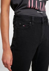 Černé vysoké džíny vyrobené z denimu, které mají přední kapsu s malou vyšívanou štítkovou cedulkou a klasické zapínání na knoflík a zip.