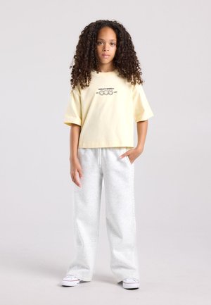 Fille aux cheveux bouclés portant un t-shirt oversize jaune clair et un pantalon de survêtement gris, debout devant un fond uni.
