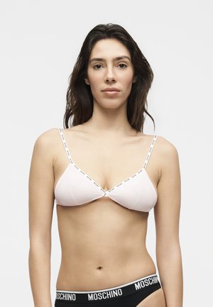 Kobieta o długich ciemnych włosach, ubrana w biały biustonosz z cienkimi ramiączkami z logo i czarne majtki marki Moschino, stojąca na jednolitym tle.