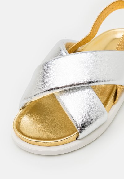 Sandal in metallo argentato con cinturini incrociati, interno dorato e suola bianca. Presenta una texture liscia e un design con tacco basso.