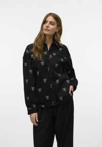 Zwarte shirt met witte strikpatronen, gemaakt van een zachte stof, met een kraag en lange mouwen, sluiting met knopen aan de voorkant en zakken.