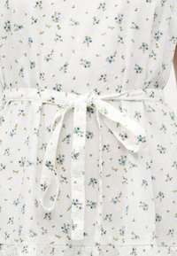 Λευκό ύφασμα με μικρό μπλε και πράσινο ανθικό μοτίβο, διαθέτει ζώνη στη μέση δεμένη και διακριτικές υφασμάτινες κουκκίδες.