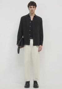 Junger Mann steht, trägt ein schwarzes Hemd über einem schwarzen Oberteil, beigen Hosen, schwarzen Schuhen und hält eine gefaltete schwarze Tasche vor einem schlichten weißen Hintergrund.