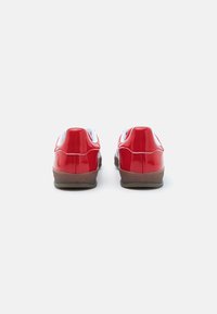 Red patent leather shoes with white accents, rounded shape, flat sole, and contrasting brown outsole. Simple, clean design with no embellishments.