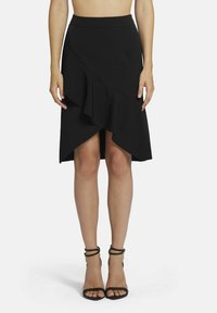 Zwarte wikkelrok met asymmetrische zoom, voorzien van een ruchedetail. Gemaakt van een soepele stof. Gecombineerd met zwarte sandalen met hoge hakken en bandjes.