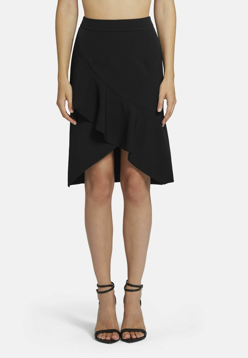 Zwarte wikkelrok met asymmetrische zoom, voorzien van een ruchedetail. Gemaakt van een soepele stof. Gecombineerd met zwarte sandalen met hoge hakken en bandjes.