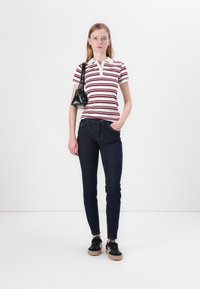 Νεαρή γυναίκα φορώντας ένα λευκό και μπορντό ριγέ πόλο, σκούρα skinny τζιν, μαύρα sneakers και κρατώντας μια μαύρη τσάντα ώμου.