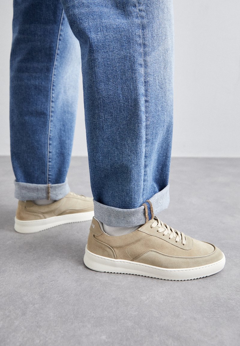 Beige suede sneakers with white rubber soles, featuring tonal laces and subtle side stitching, paired with rolled-up light blue denim jeans.