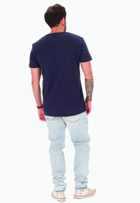 T-shirt blu navy a maniche corte con scollo rotondo, abbinata a jeans azzurri e scarpe da ginnastica beige. Tatuaggi visibili sulle braccia.