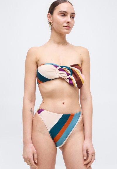 Vrouw draagt een strapless gestreepte bikini in meerdere kleuren met een grote cirkelvormige ringdetail op het bovenstuk, staand tegen een effen lichte achtergrond.
