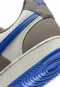 Primer plano de una zapatilla con una parte superior en beige y blanca, con una franja azul y una suela texturada, destacando el logo de la marca y los detalles de costura.