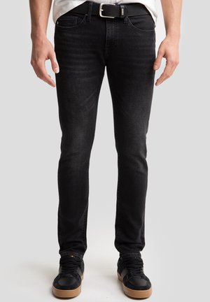 Jeans in denim neri con vestibilità slim, caratterizzati da un lavaggio sbiadito, design a cinque tasche e dettagli cuciti. Indossati con una cintura nera e sneaker.
