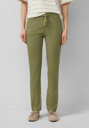 Vrouw met olijfgroene broek met rechte pijpen en trekkoord in de taille, beige casual schoenen en een licht gestreept shirt, staand tegen een eenvoudige grijze muur.