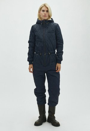 Femme portant une combinaison matelassée bleu marine à capuche avec poches avant et bottes noires mi-mollet, debout devant un fond clair uni.
