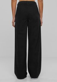 Persona con pantalones negros de cintura alta y pata ancha, de pie, vista desde atrás sobre un fondo gris liso.