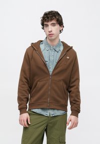 Καφέ φούτερ με φερμουάρ από μαλακό ύφασμα, με μπροστινή τσέπη και ρυθμιζόμενο κουκούλα, φορεμένο πάνω από ένα ανοιχτό μπλε τζιν πουκάμισο και πράσινο παντελόνι.