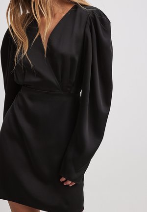 Vrouw draagt een zwarte satijnen jurk met lange pofmouwen en een V-hals, staand tegen een effen lichte achtergrond.