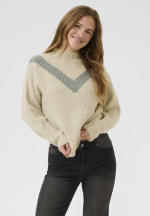 Cream-colored ribbed sweater with a high neck and a gray chevron pattern on the chest, paired with dark jeans. Relaxed fit and textured fabric.