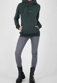 Sudadera con capucha de color verde oscuro con bolsillo tipo canguro, cierre con cordón, ajuste casual, combinada con jeans pitillo grises y botines negros.