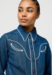 Donna con capelli intrecciati che indossa una camicia di denim blu scuro con piping bianco e dettagli a freccia sul petto, che guarda di lato.