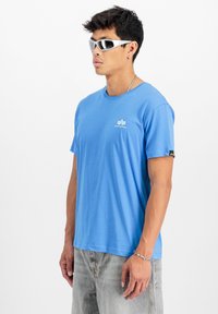 Zila kokvilnas T-kreklu ar apaļo kakla izgriezumu, īsām piedurknēm un mazu baltu logotipu uz krūtīm. Saskaņojot ar gaiši pelēkiem džinsu šortiem.